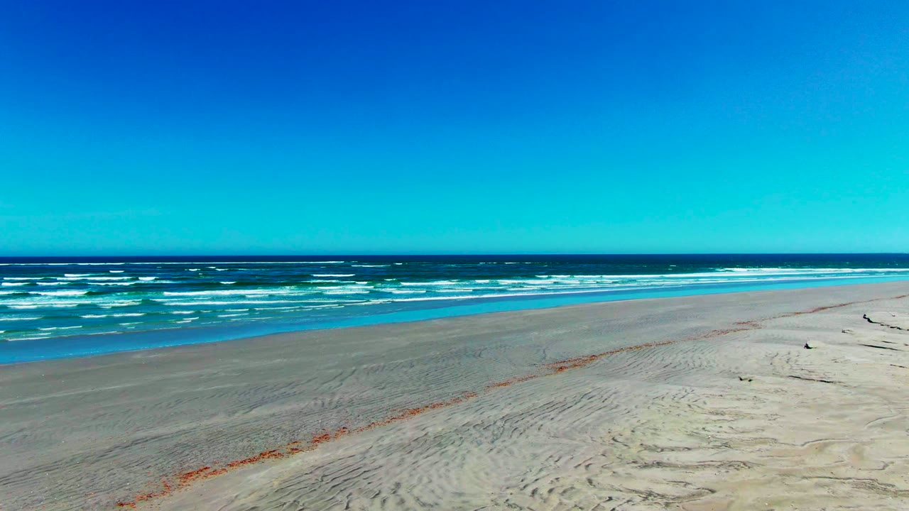 La Bocana shoreline seen from above, with a broad strip of empty sand, scattered drift material near the beach line, blue-green water, whitecaps, and no visible people.