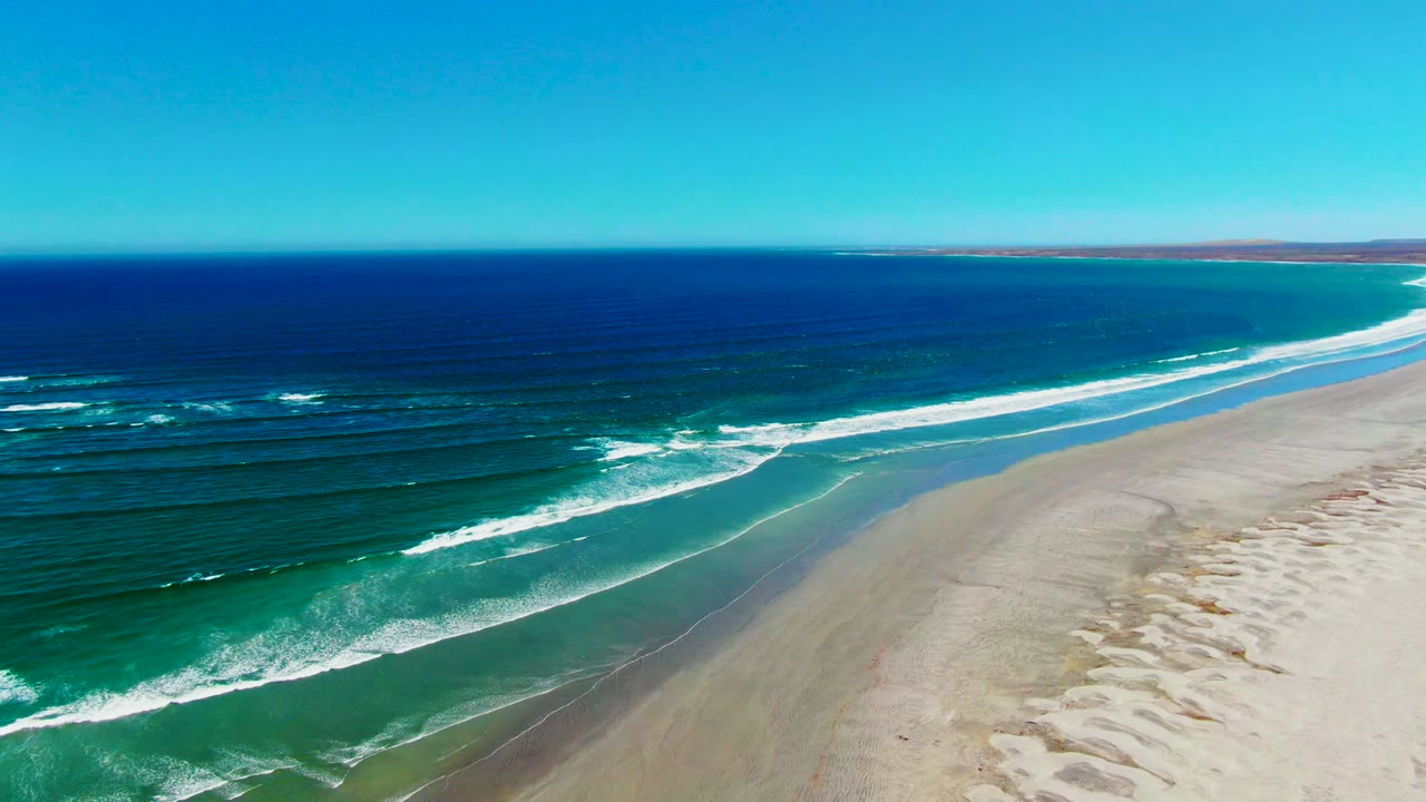 Aerial frame over La Bocana, Baja California Sur showing deep blue ocean, turquoise surf, pale sand, distant low coast, and no visible people.