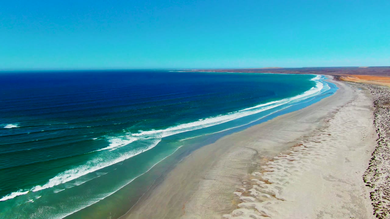 High aerial view over La Bocana, Baja California Sur, with deep blue ocean on the left, white surf approaching the sandy beach, tan land on the right, and no visible people.