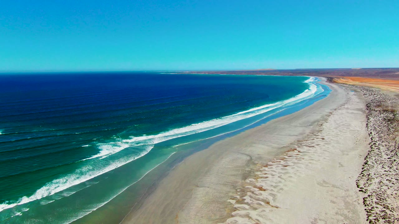 Aerial coastline frame at La Bocana showing a long white wave line closer to shore, turquoise shallows beside pale sand, arid land beyond the beach, and no visible people.