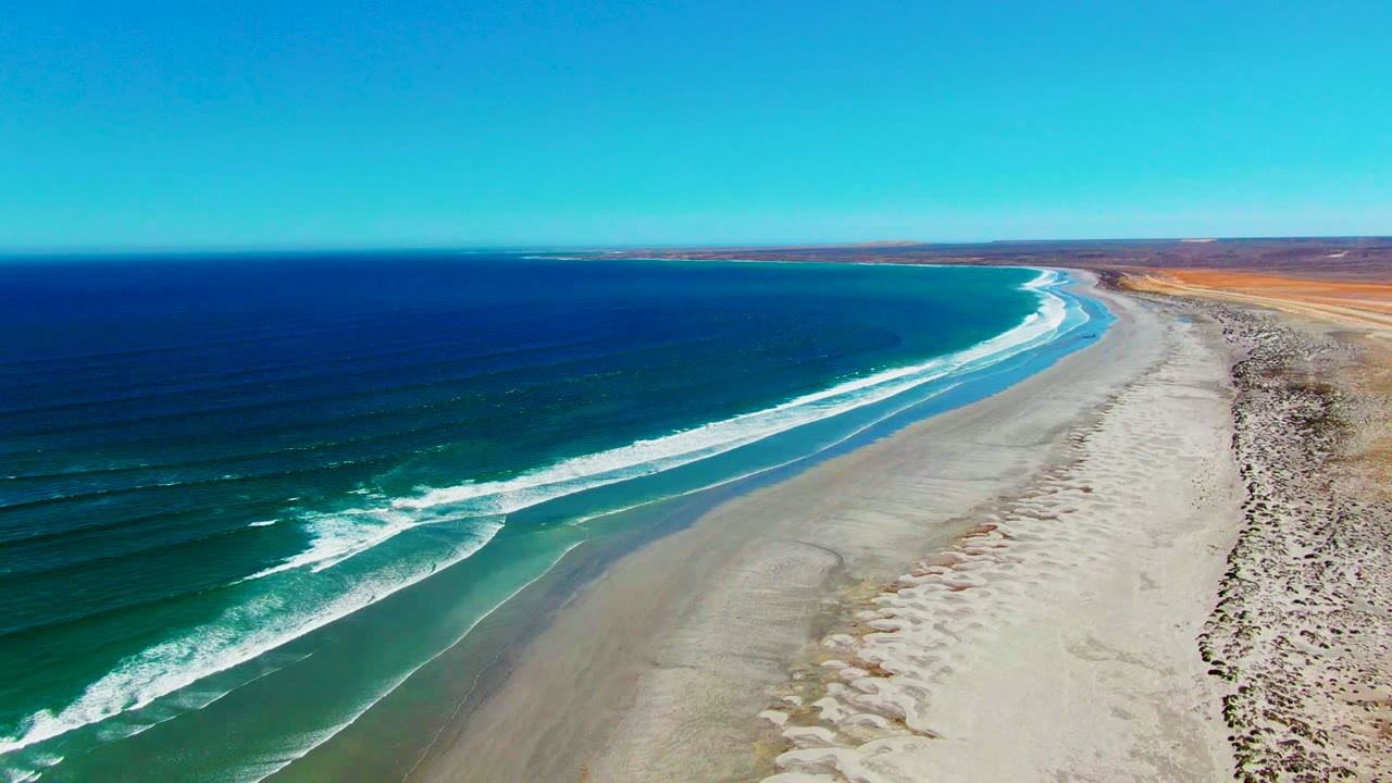 Drone-style view of the La Bocana shoreline with the beach curving toward the horizon, dark blue water offshore, bright foam bands near the sand, and no visible people.