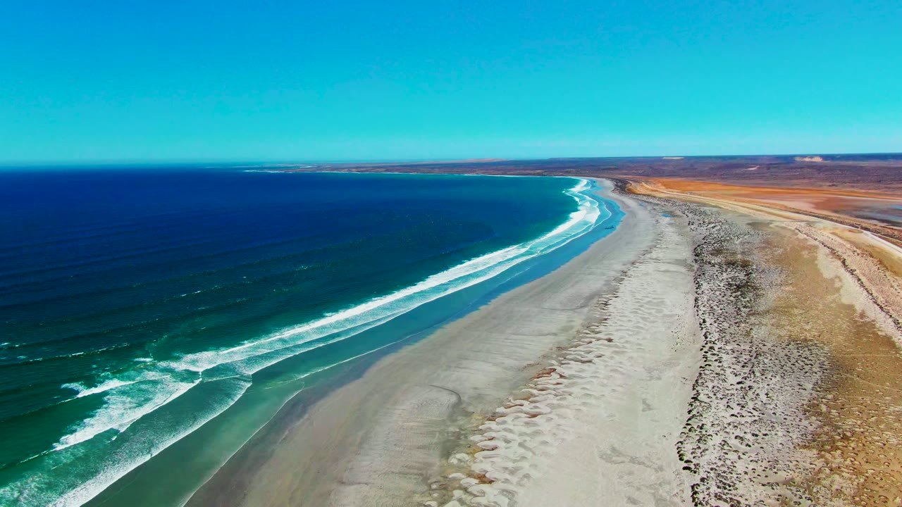 At 0:00, an aerial view over La Bocana, Baja California Sur shows blue ocean, white surf, pale sand, dry shoreline scrub, a clear sky, and no visible people.