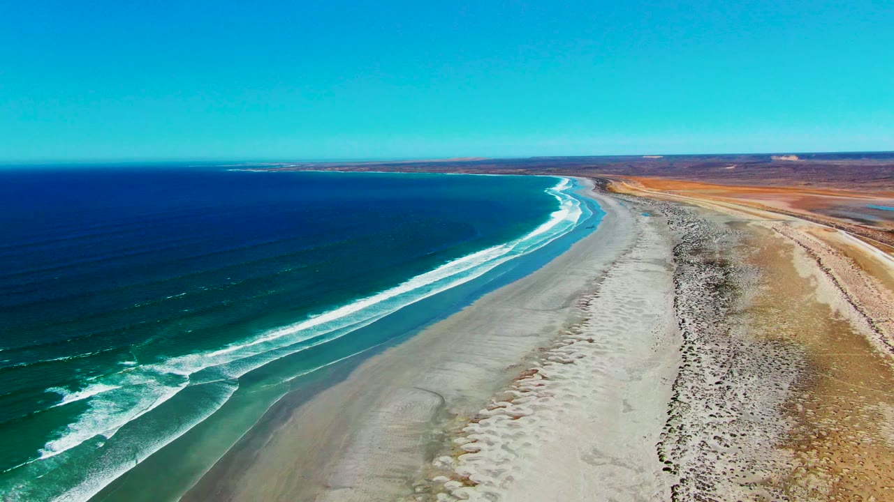 Drone view of curved surf lines reaching a wide sandbar and dry shoreline.