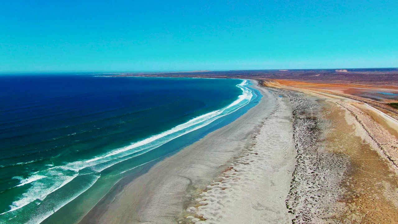 At 0:02, white foam traces the edge of the beach at La Bocana while blue water fills the left side, dry coastal terrain fills the right side, and no visible people appear.