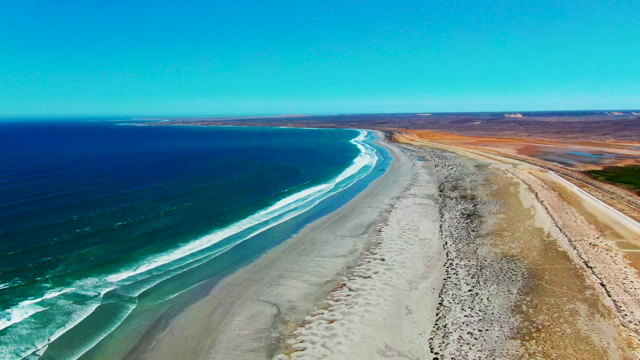 Aerial view over La Bocana, Baja California Sur, with deep blue ocean on the left, white surf curving onto a wide sandy shore, dry rocky land on the right, and no visible people.