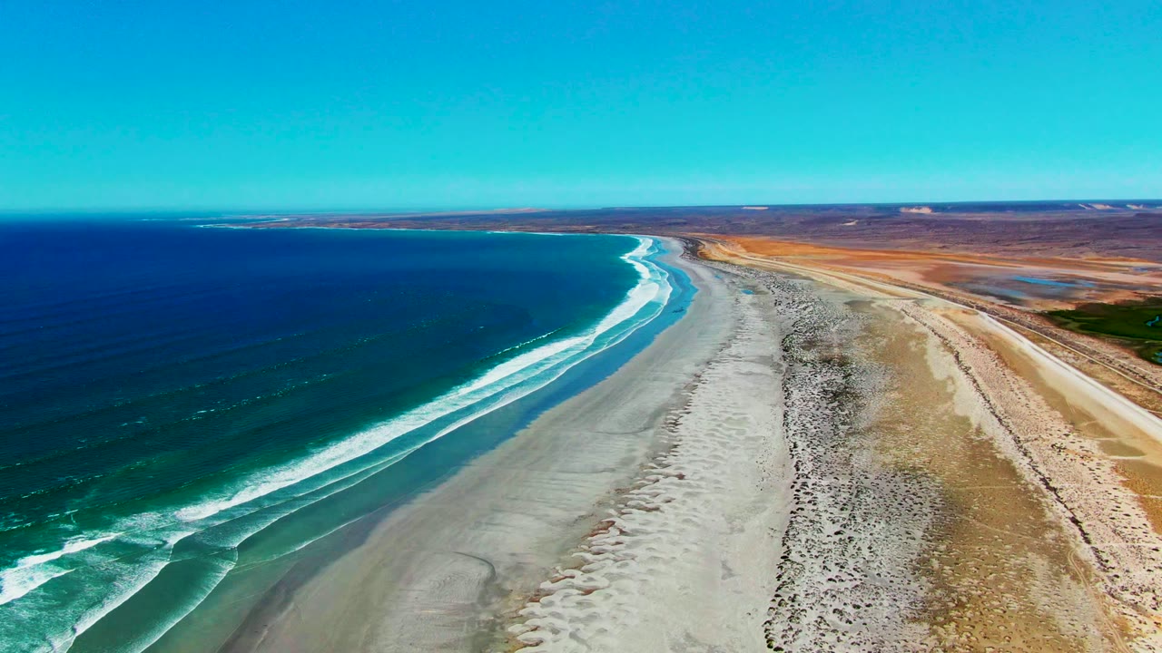 White waves curl along a long sandy beach beside dry coastal land.