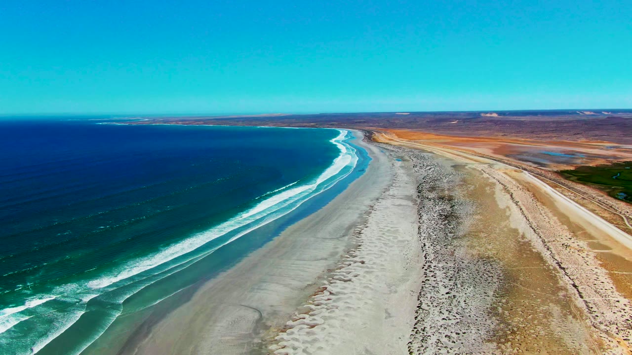 Aerial view of the La Bocana coastline with turquoise nearshore water, white foam tracing the beach edge, pale sand in the center, dry terrain to the right, and no visible people.
