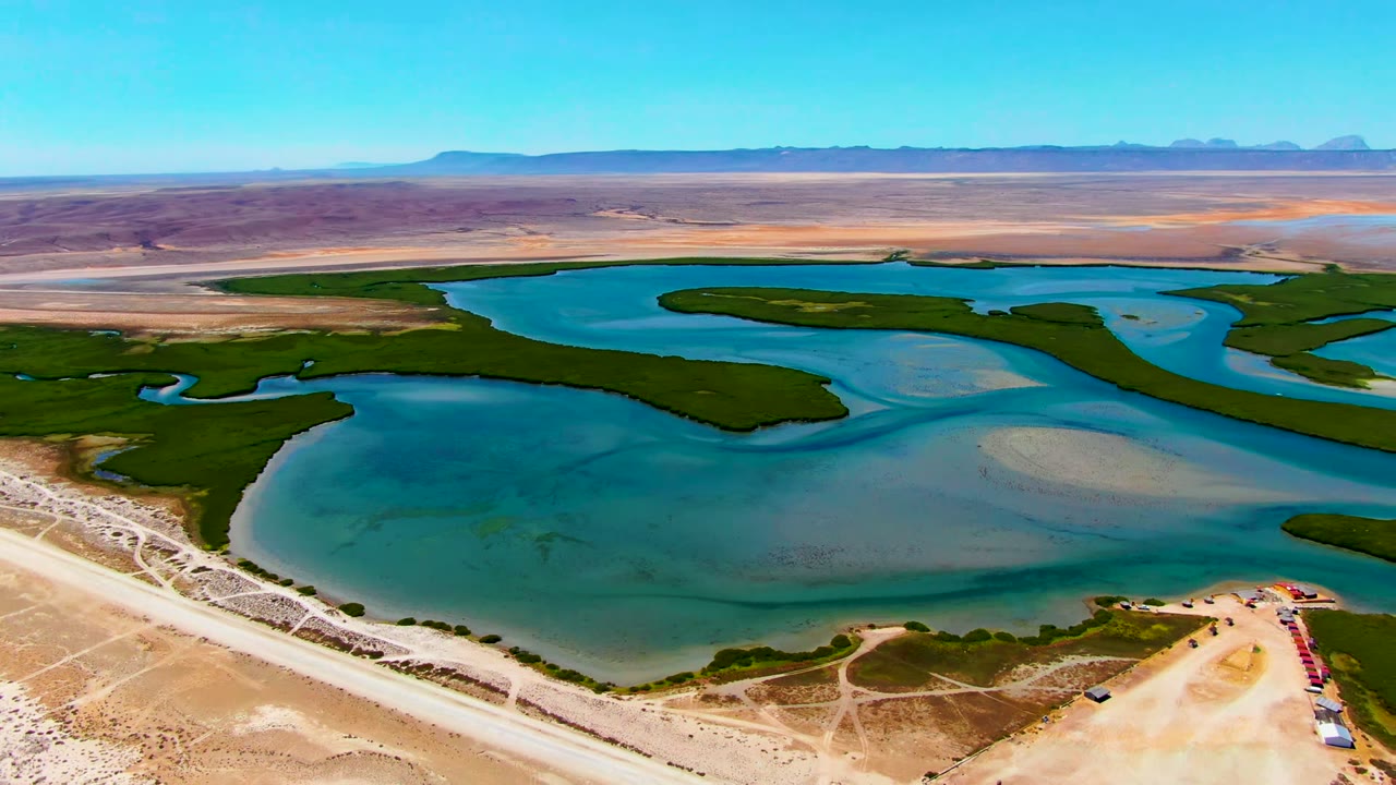 Opening aerial frame over a desert reservoir near La Bocana, Baja California Sur, with green vegetation bands, a pale dirt road, tan arid ground, distant mountains, and no visible people.