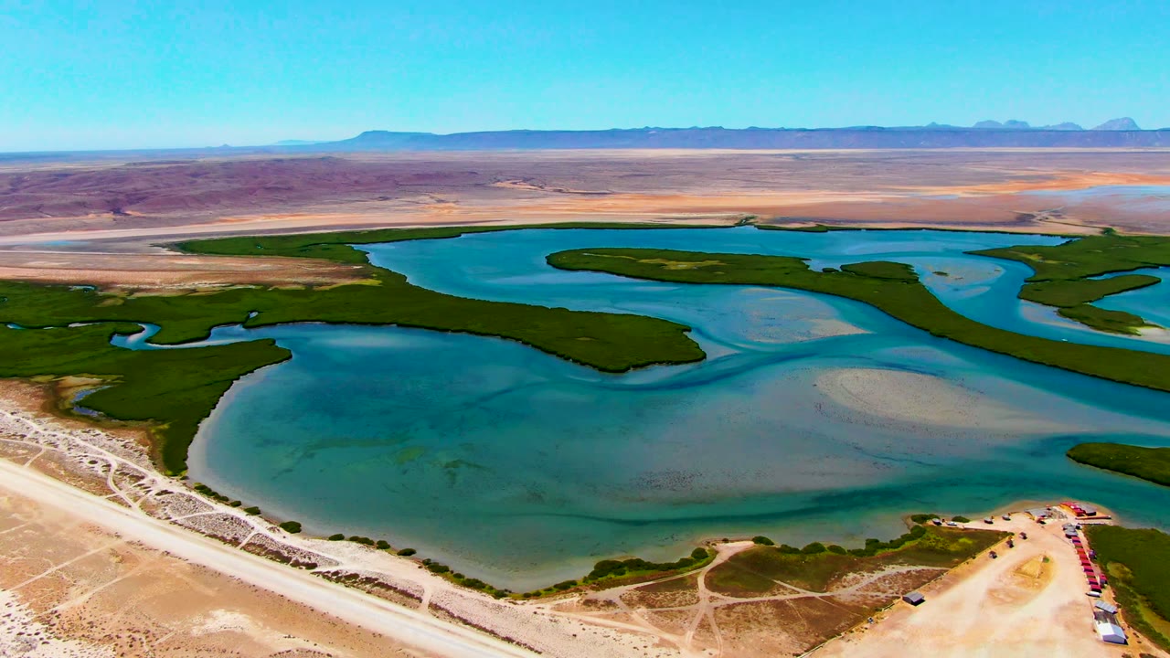 Wide drone frame near La Bocana, Baja California Sur with shallow blue water, curved green banks, pale sandbars, a dirt road along the lower edge, desert background, and no visible people.