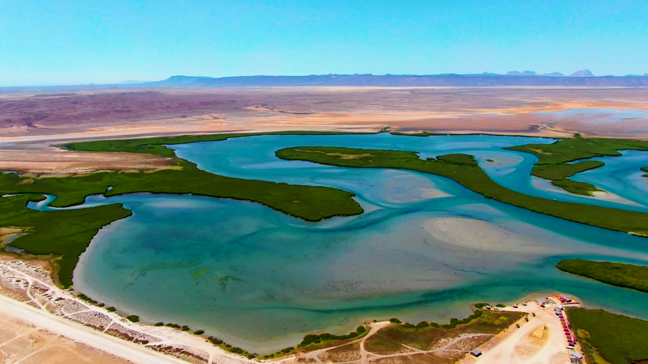 Opening aerial frame over La Bocana, Baja California Sur showing blue-green reservoir water, dark vegetation bands, pale desert terrain, distant mountains, and no visible people.