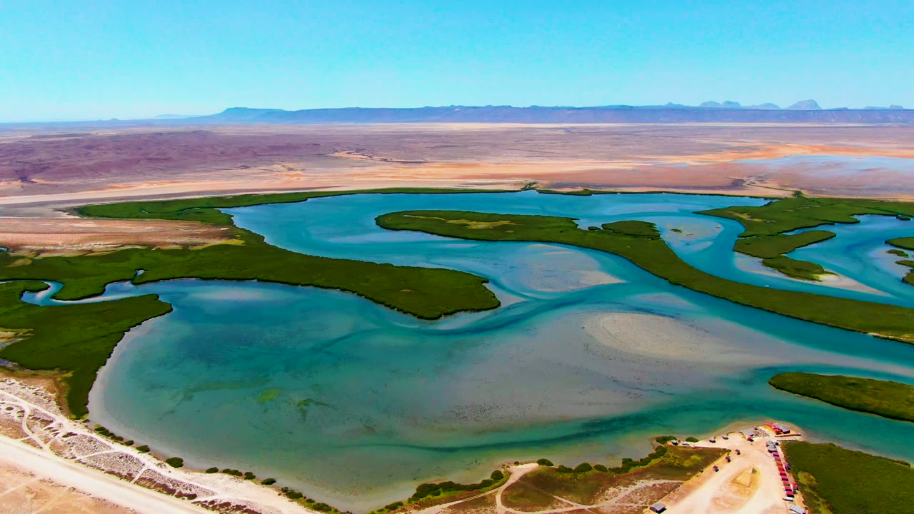Aerial frame at 0:02 near La Bocana showing curving blue water, dark green channel edges, pale sand flats, a small shoreline access area, distant mountains, and no visible people.