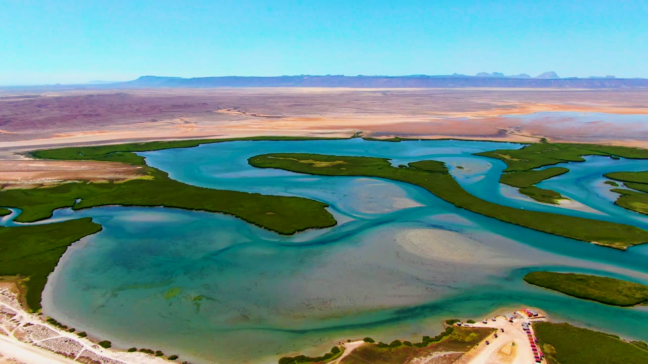 Aerial view near La Bocana, Baja California Sur, showing turquoise water channels, green wetland strips, pale sandbars, distant arid land, and no visible people.