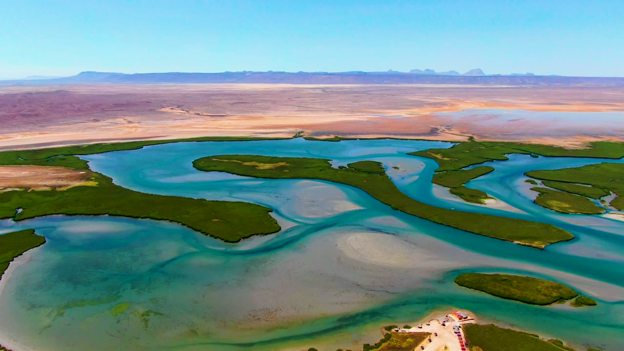 Aerial frame over La Bocana, Baja California Sur showing turquoise channels around green islands, pale sandbars, distant desert mountains, a small shoreline structure at the lower edge, and no visible people.
