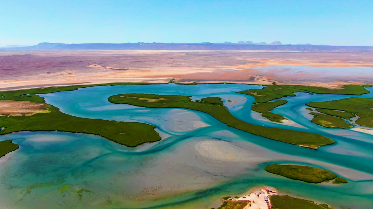 Aerial frame over La Bocana, Baja California Sur showing shallow turquoise water divided by green islands and sandbars, with open arid land and mountains under a clear sky, and no visible people.