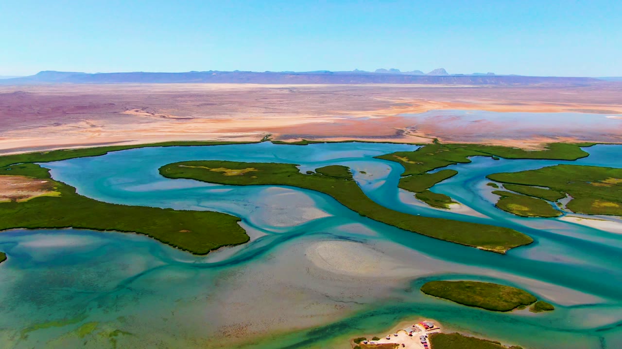 Opening aerial frame over La Bocana, Baja California Sur, showing turquoise water channels around green islands, pale sandbars, distant desert, and no visible people.
