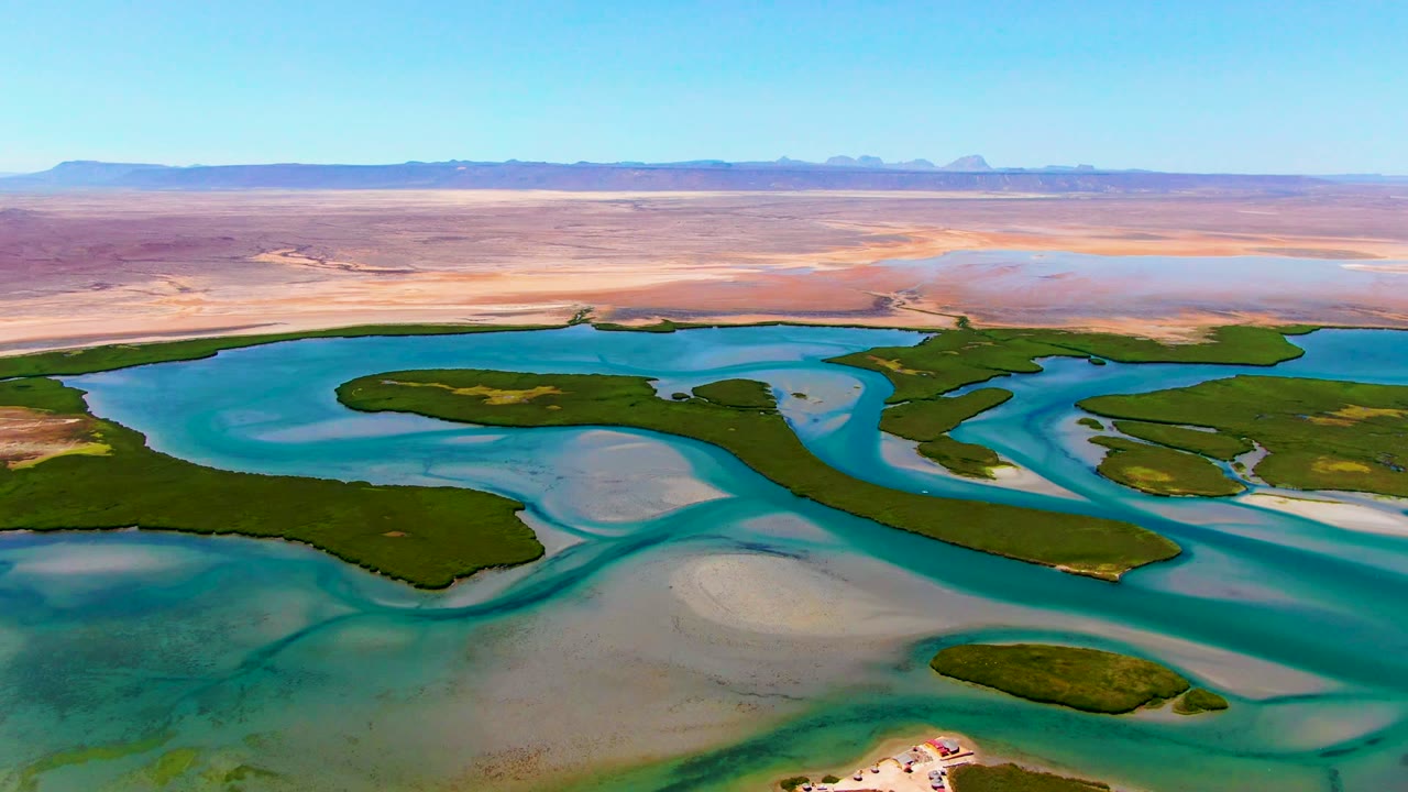 Aerial view at La Bocana, Baja California Sur, showing turquoise lagoon channels, dark green islands, pale underwater sand, arid land, and no visible people.