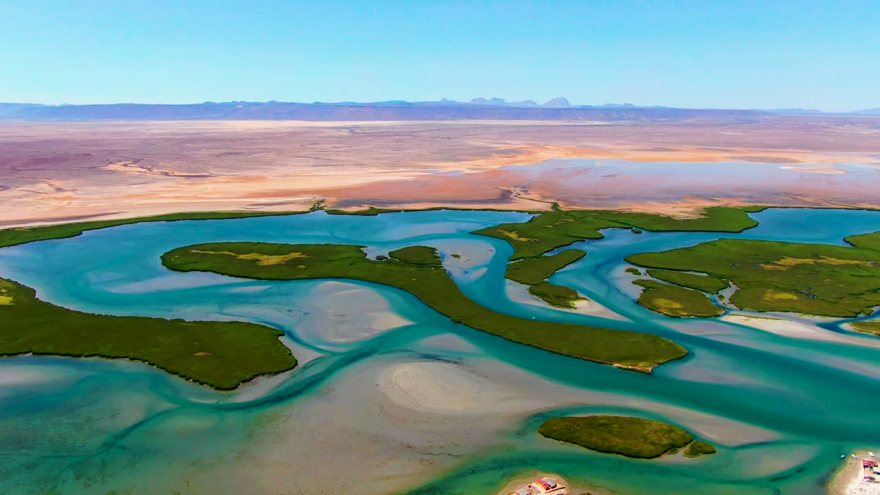 Wide aerial view over La Bocana, Baja California Sur, with turquoise channels around green islets, pale sandbars, desert flats, distant low mountains, and no visible people.