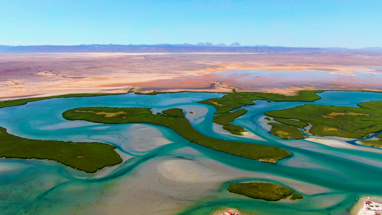 Wide drone view at La Bocana with blue-green water channels, dark green islets, light sand shoals, a dry desert plain, low mountains on the horizon, and no visible people.