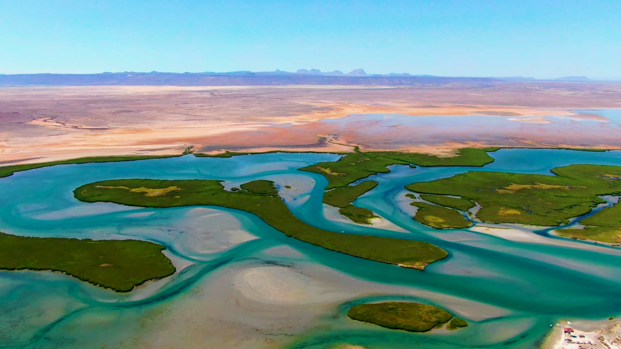 Opening aerial view over La Bocana, Baja California Sur, Mexico, with blue-green channels around green islands, pale sandbars, desert flats, distant mountains, and no visible people.