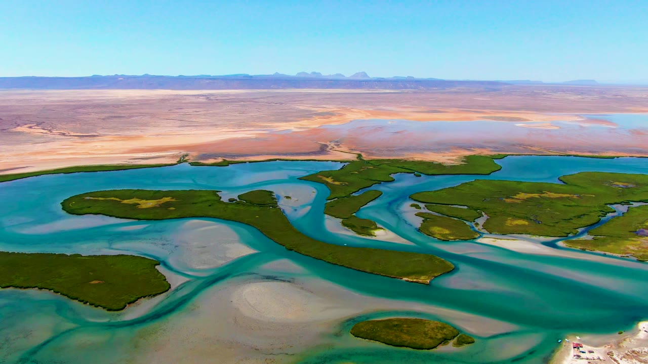 A wide La Bocana drone frame holds blue water and green wetland patches in the lower half, with broad tan desert and a clear sky across the horizon, and no visible people.