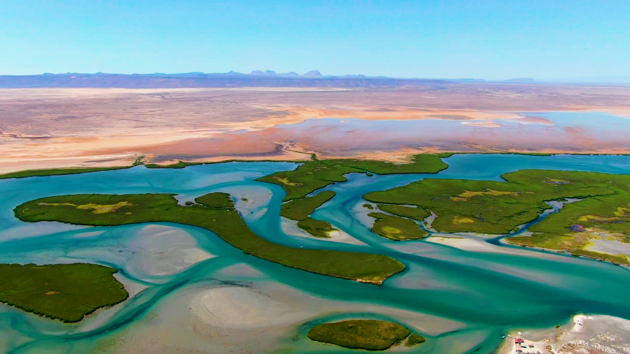 Aerial view over La Bocana, Baja California Sur, with turquoise river channels wrapping around green delta islands, pale sandbars, distant desert flats, and no visible people.