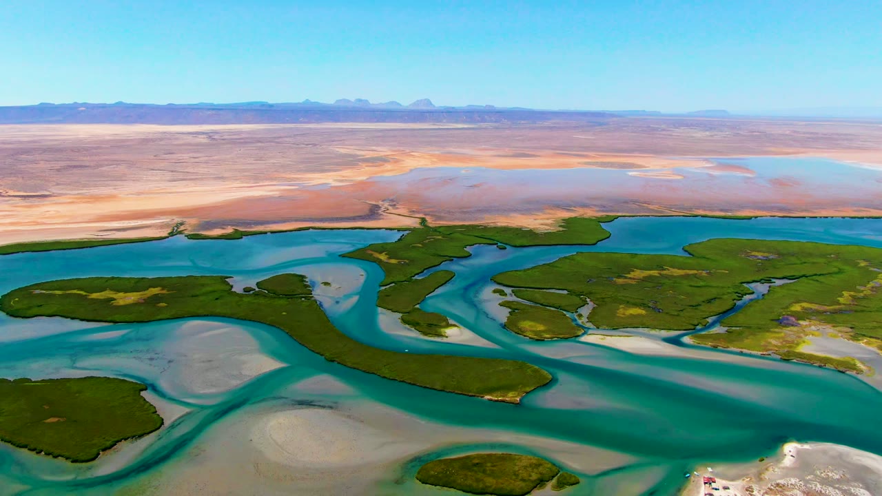Aerial view of the La Bocana delta with a broad turquoise channel, dark green island vegetation, pale submerged sand, dry desert plain, and no visible people.