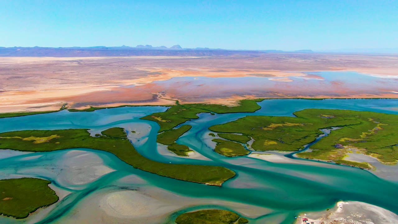 Aerial frame over La Bocana, Baja California Sur showing turquoise channels wrapping green landmasses, pale sandbars in front, tan flats beyond, distant mountains, and no visible people.