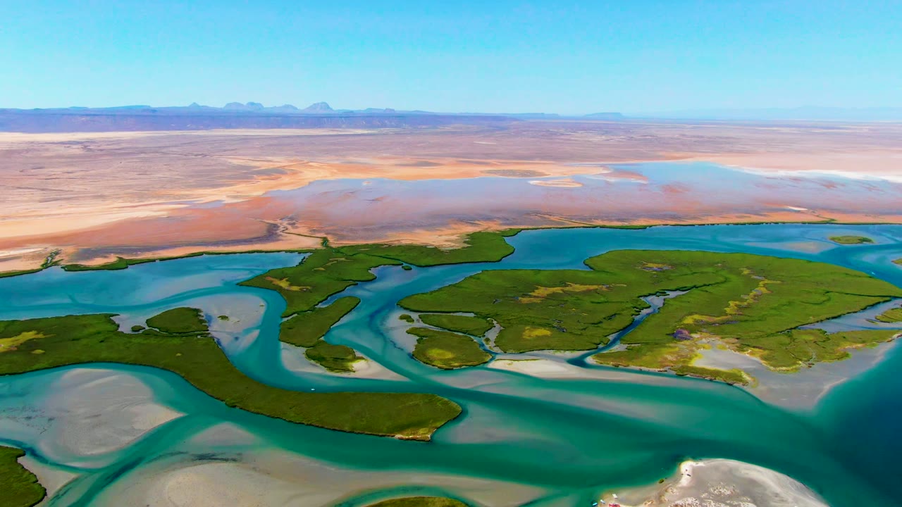 Aerial frame at La Bocana, Baja California Sur showing turquoise water curving around green islands with pale sand bars, pink-tan desert flats, blue sky, and no visible people.