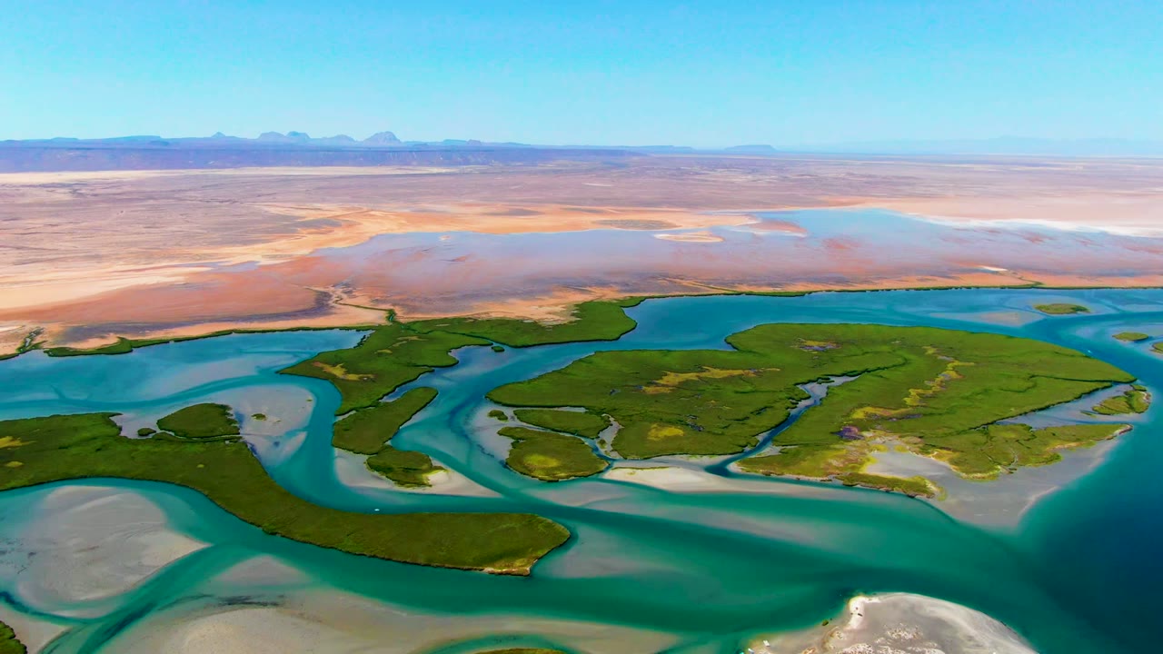 Aerial frame at La Bocana, Baja California Sur showing a broad turquoise channel at right, darker green islands across the center, pale shallows at left, desert flats, and no visible people.