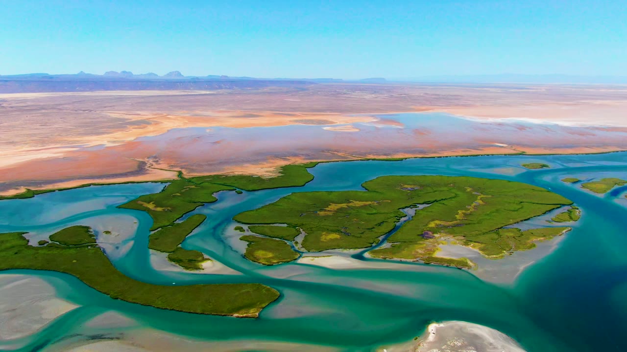 Opening aerial frame over La Bocana, Baja California Sur, showing green delta islands divided by blue water channels, pale sandbars, desert flats, a clear sky, and no visible people.