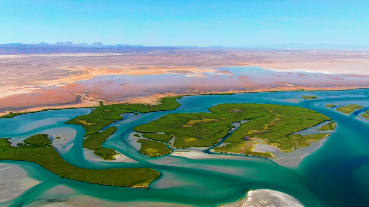 Wide aerial of turquoise waterways curving around green islands beside a sandy desert plain.