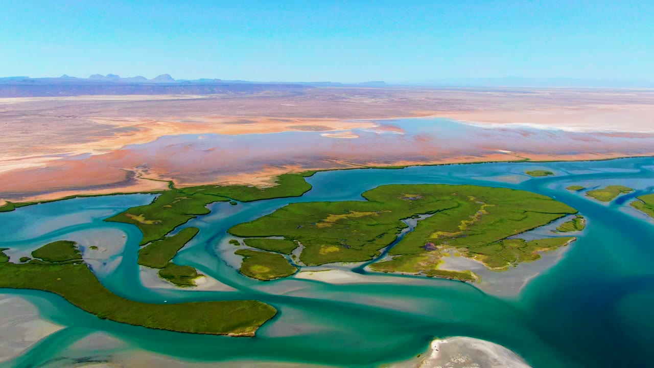 Wide drone-height frame over La Bocana, Baja California Sur, showing blue coastal water around green islands, exposed sandbars, a broad desert background, mountain silhouettes, and no visible people.