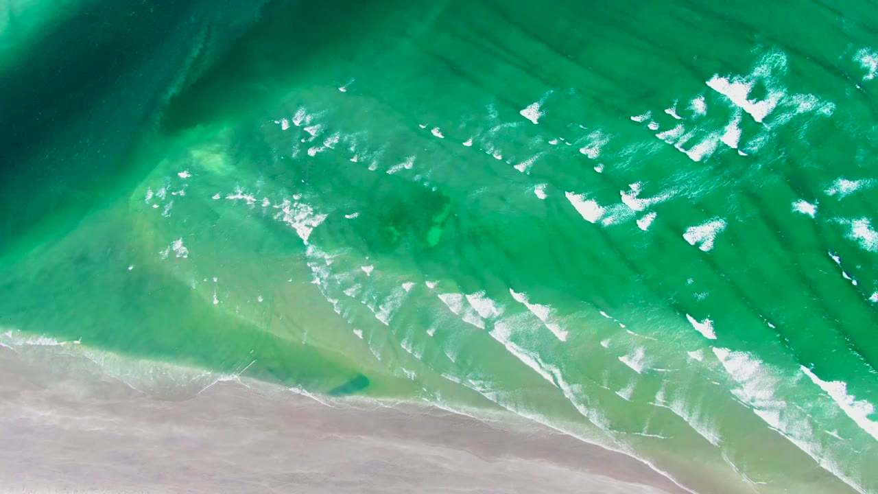 Overhead view in La Bocana, Baja California Sur, with green-turquoise water, white foam bands, pale sand along the lower edge, and no visible people.