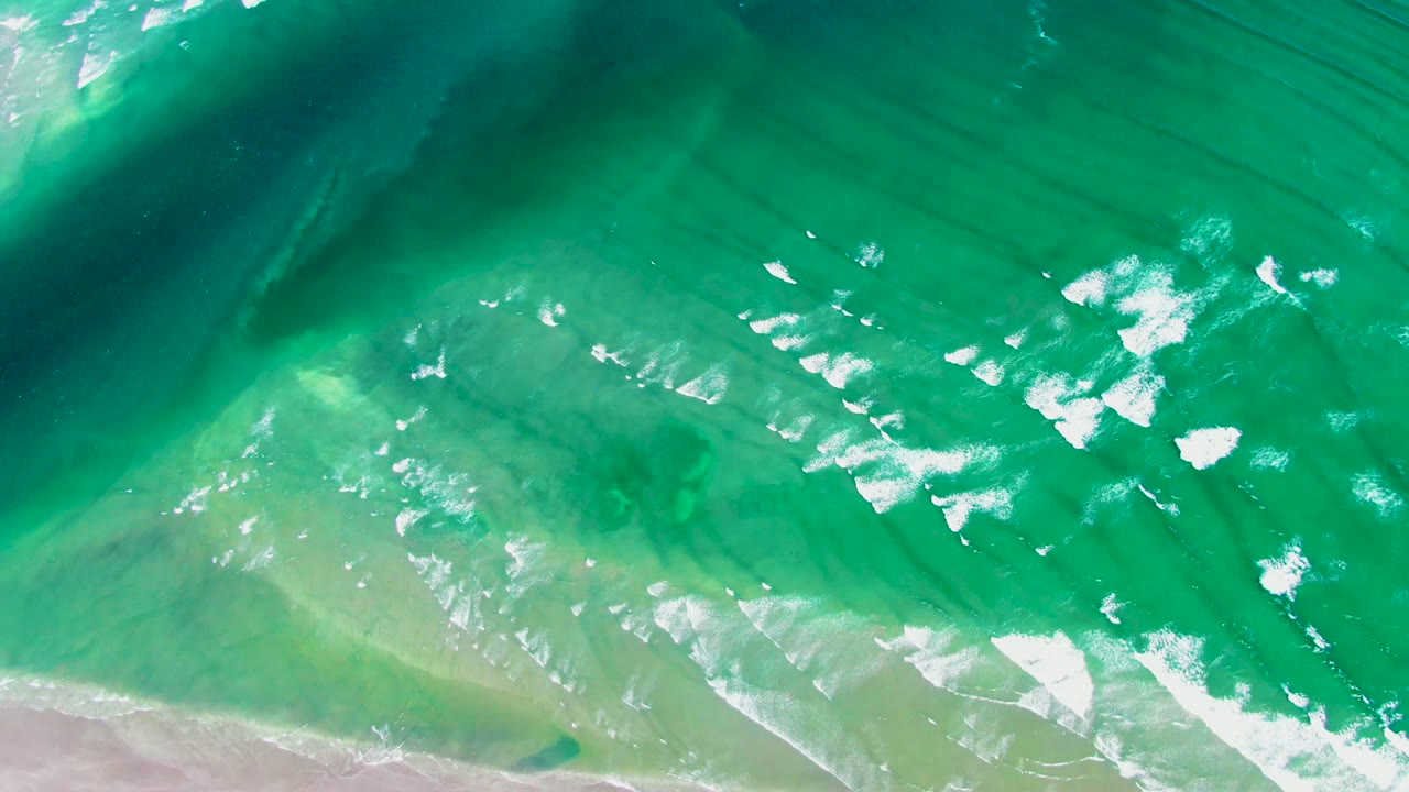 At 0:02, the aerial view over La Bocana keeps the beach along the bottom while white foam clusters sit farther into the green-blue water, with no visible people.