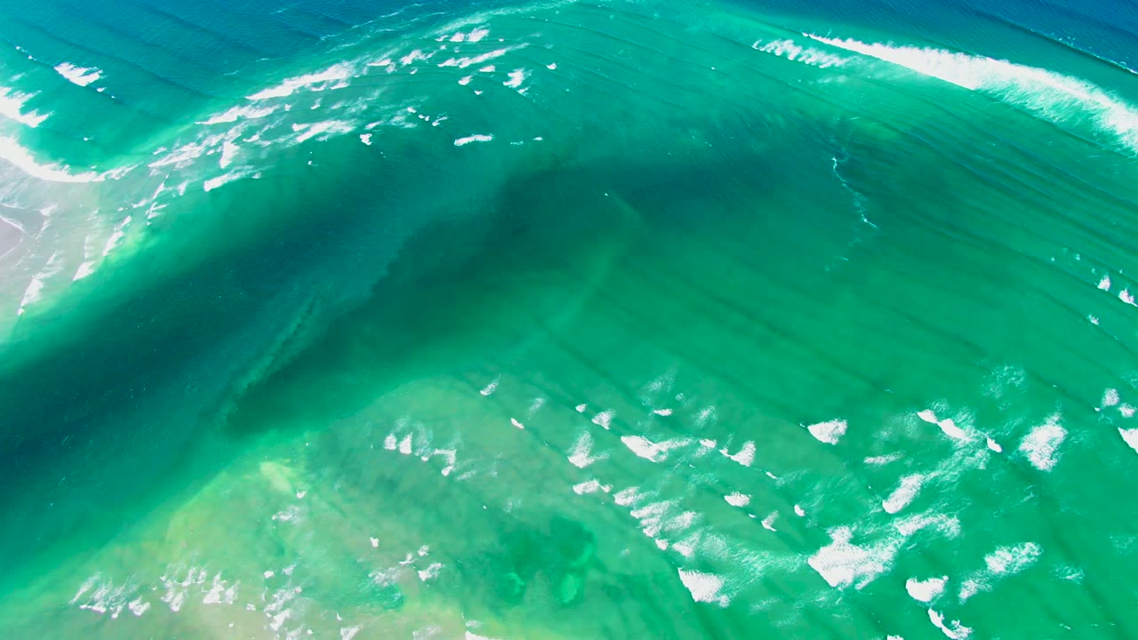 Aerial view of a turquoise wave beginning to curl across clear water near La Bocana, Baja California Sur, with white foam on the crest and no visible people.
