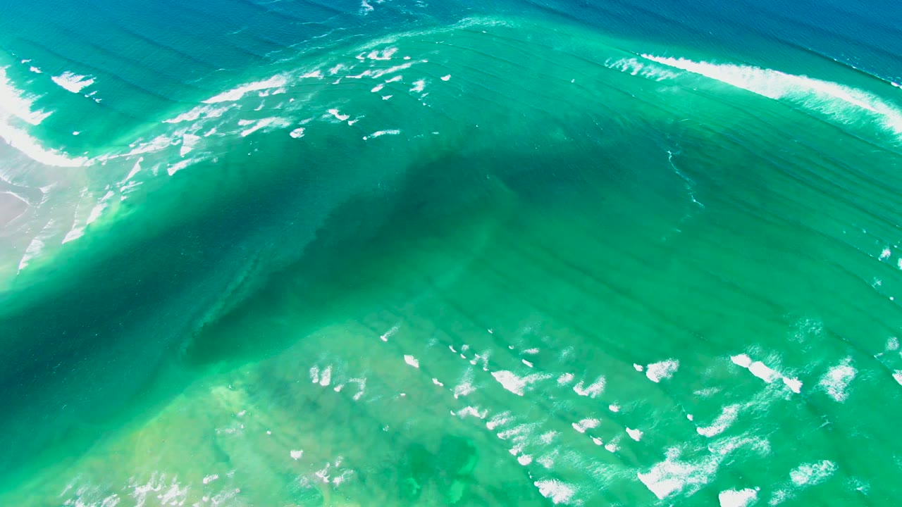 Aerial view of the curling ocean wave stretching diagonally over turquoise water near La Bocana, Baja California Sur, with foam patches scattered below and no visible people.