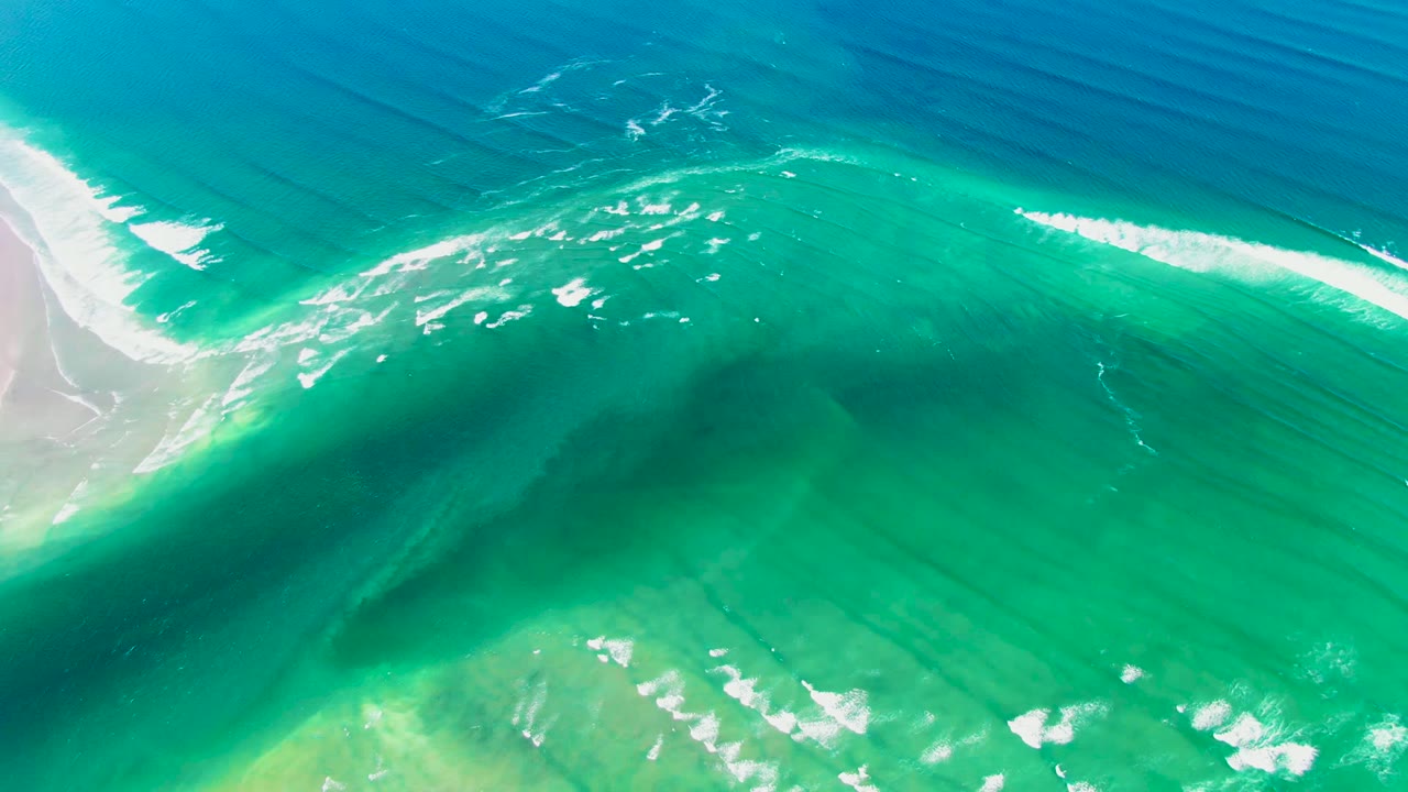 Aerial view of foamy surf over turquoise water near the pale sandy margin at La Bocana, Baja California Sur, with no visible people.