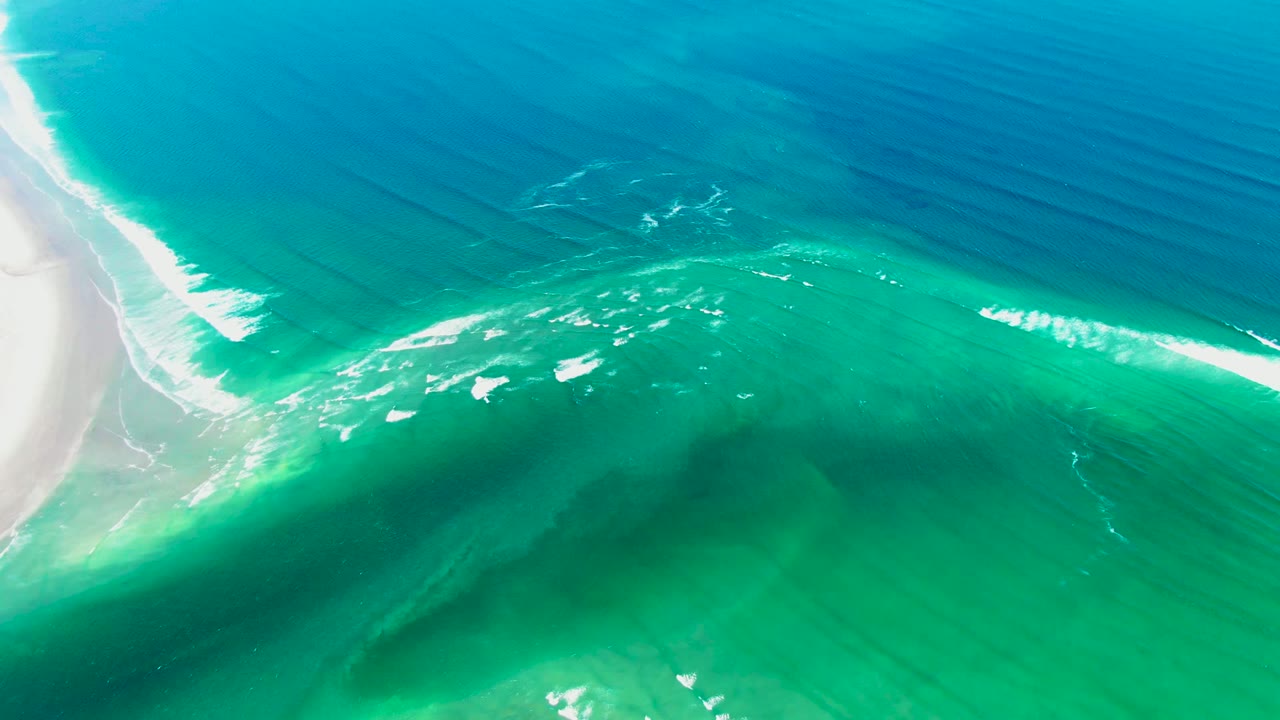 Aerial frame from La Bocana, Baja California Sur showing white sand along the left edge, turquoise shallows, blue ocean, small foam lines, and no visible people.