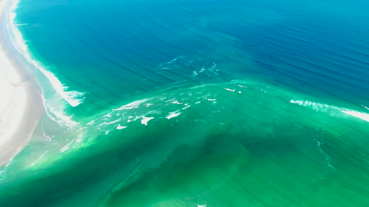 Aerial frame from La Bocana, Baja California Sur showing a broad turquoise shelf, scattered white foam patches, deep blue water across the top, a narrow strip of sand at left, and no visible people.
