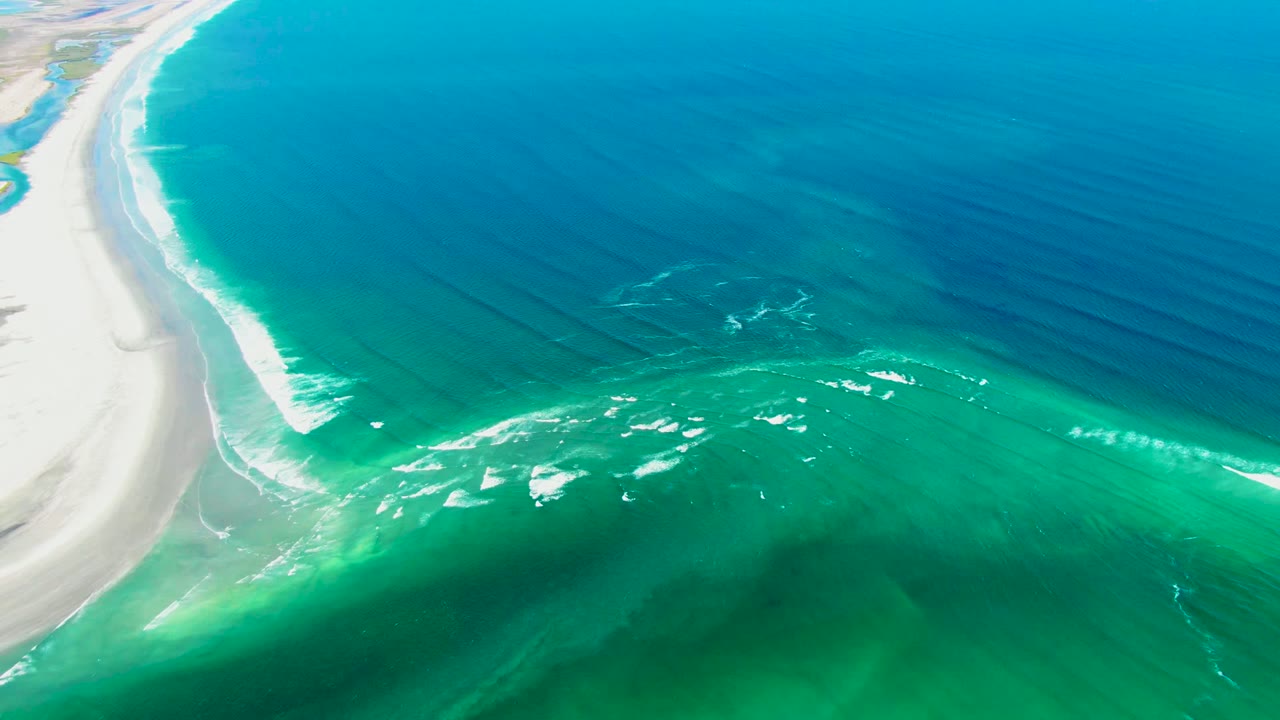Aerial view over La Bocana, Baja California Sur, with white sand curving along the left edge, turquoise shallows in the foreground, darker ocean to the right, and no visible people.