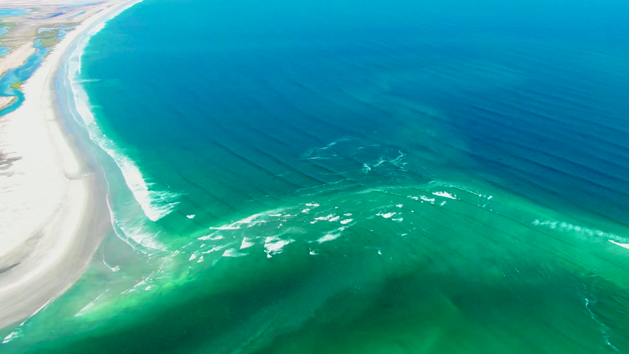 Curving shoreline with pale sand, turquoise water, and foam bands offshore.