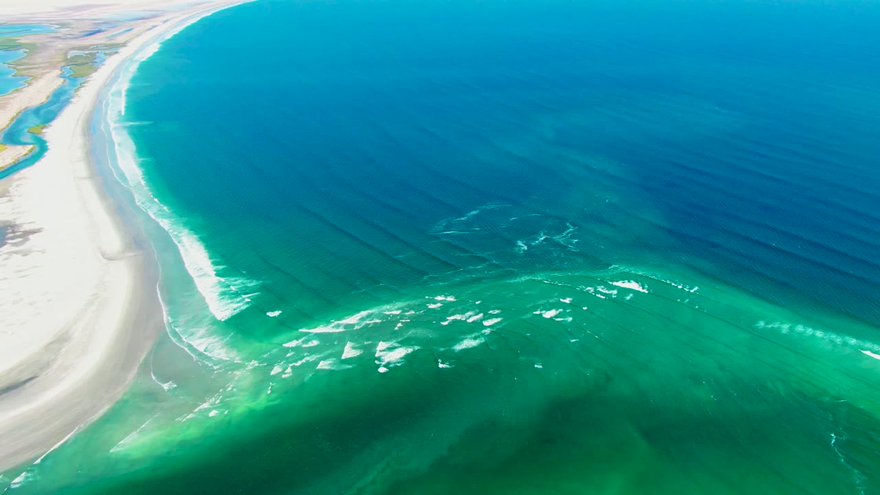 Aerial view over La Bocana showing the coastline sweeping upward along the left side, layered turquoise and blue water across the frame, bright foam over the shallows, and no visible people.