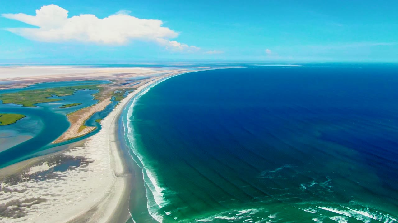 Drone view along a curved beach with ocean surf and lagoon water.