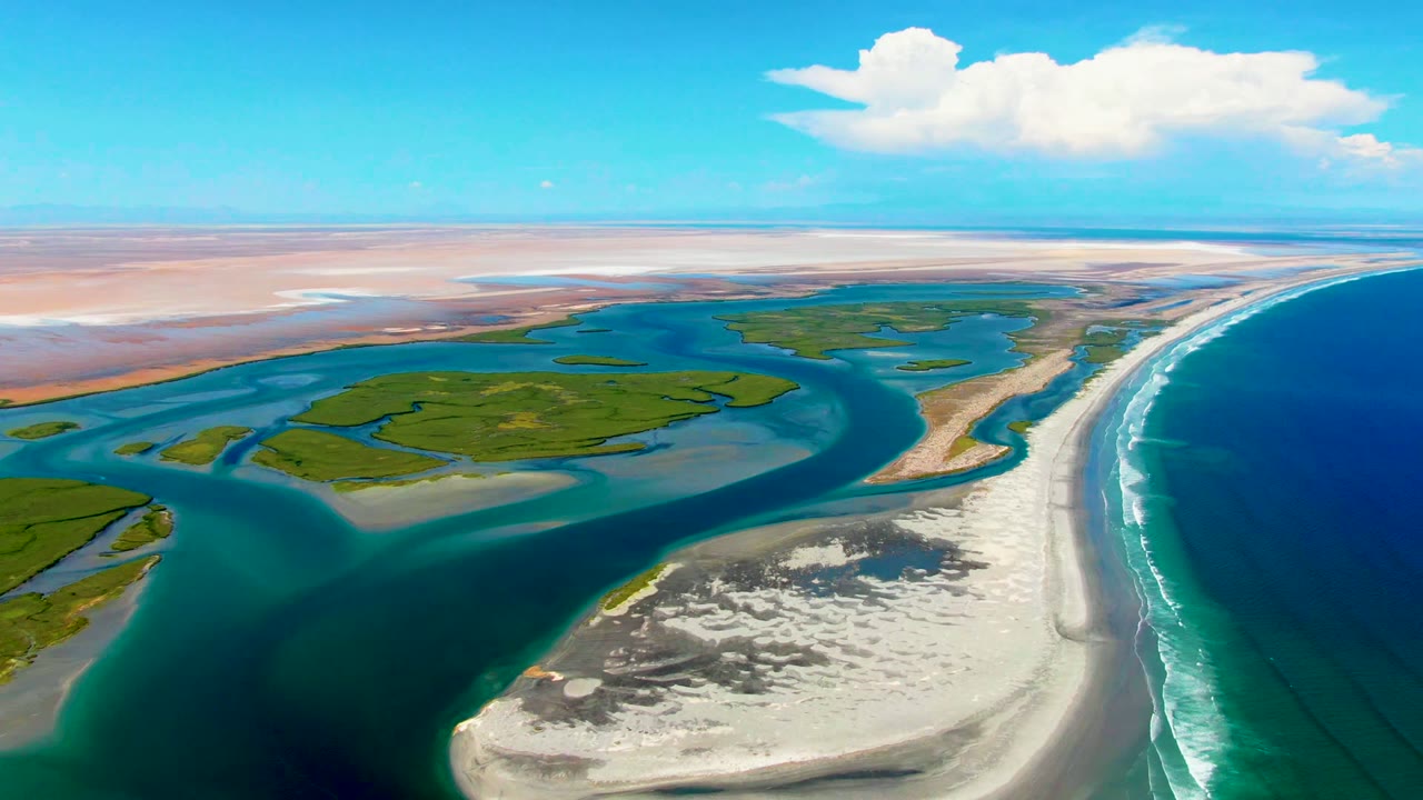 At 0:02, the La Bocana shoreline remains framed from above with green islands centered, shallow blue water around them, a long surf edge to the right, and no visible people.