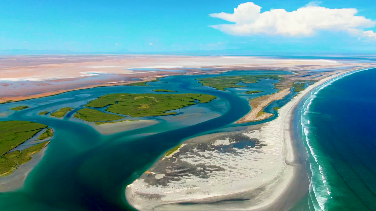 Opening aerial frame over La Bocana, Baja California Sur, showing turquoise tidal channels around green flats, pale sandbar foreground, ocean surf at right, distant tan flats, blue sky, clouds, and no visible people.