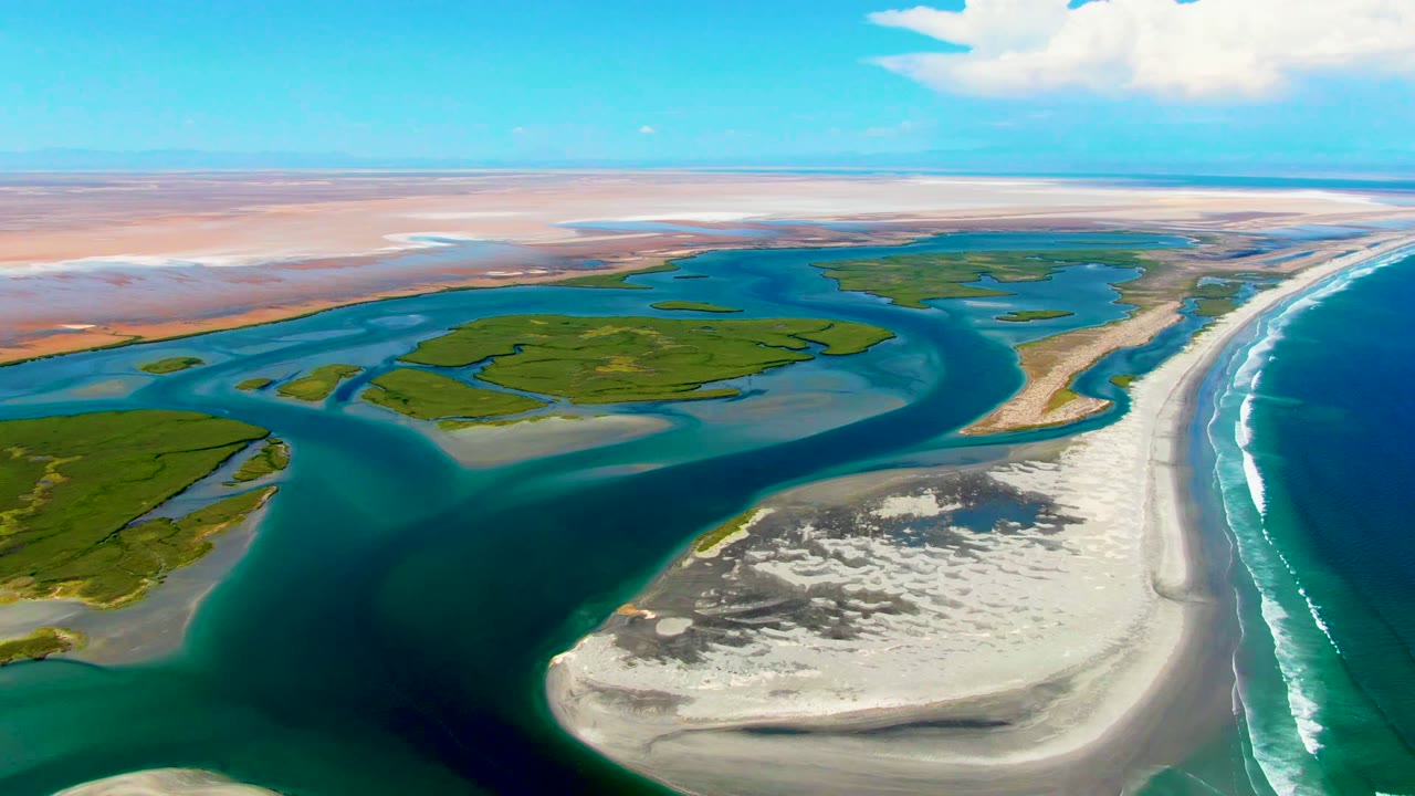 Opening aerial frame over La Bocana, Baja California Sur, showing green marsh islands divided by blue tidal channels, pale sandbars, ocean surf at right, distant tan flats, and no visible people.