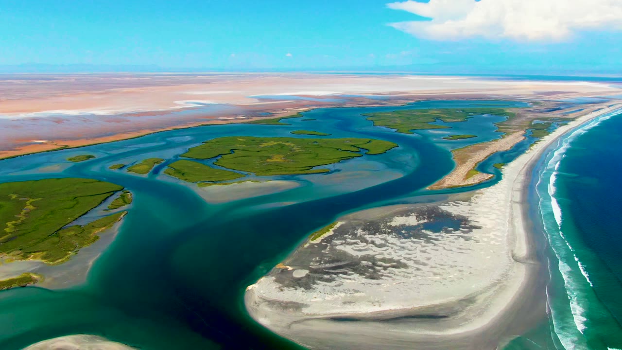 Wide drone view of tidal channels, sandbar, green marsh, and ocean surf.