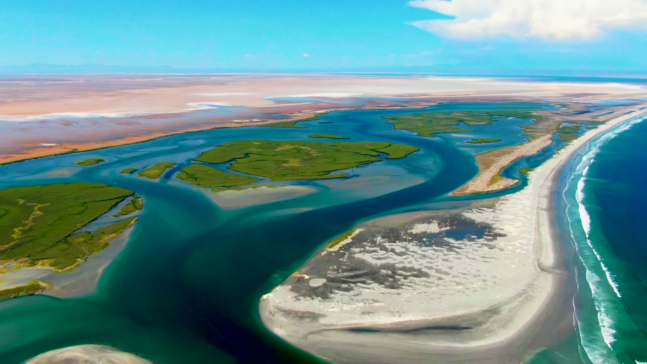 Aerial frame at 0:02 in La Bocana, Baja California Sur, with green estuary islands centered between blue water channels, pale beach sand to the right, breaking waves, bright sky, and no visible people.