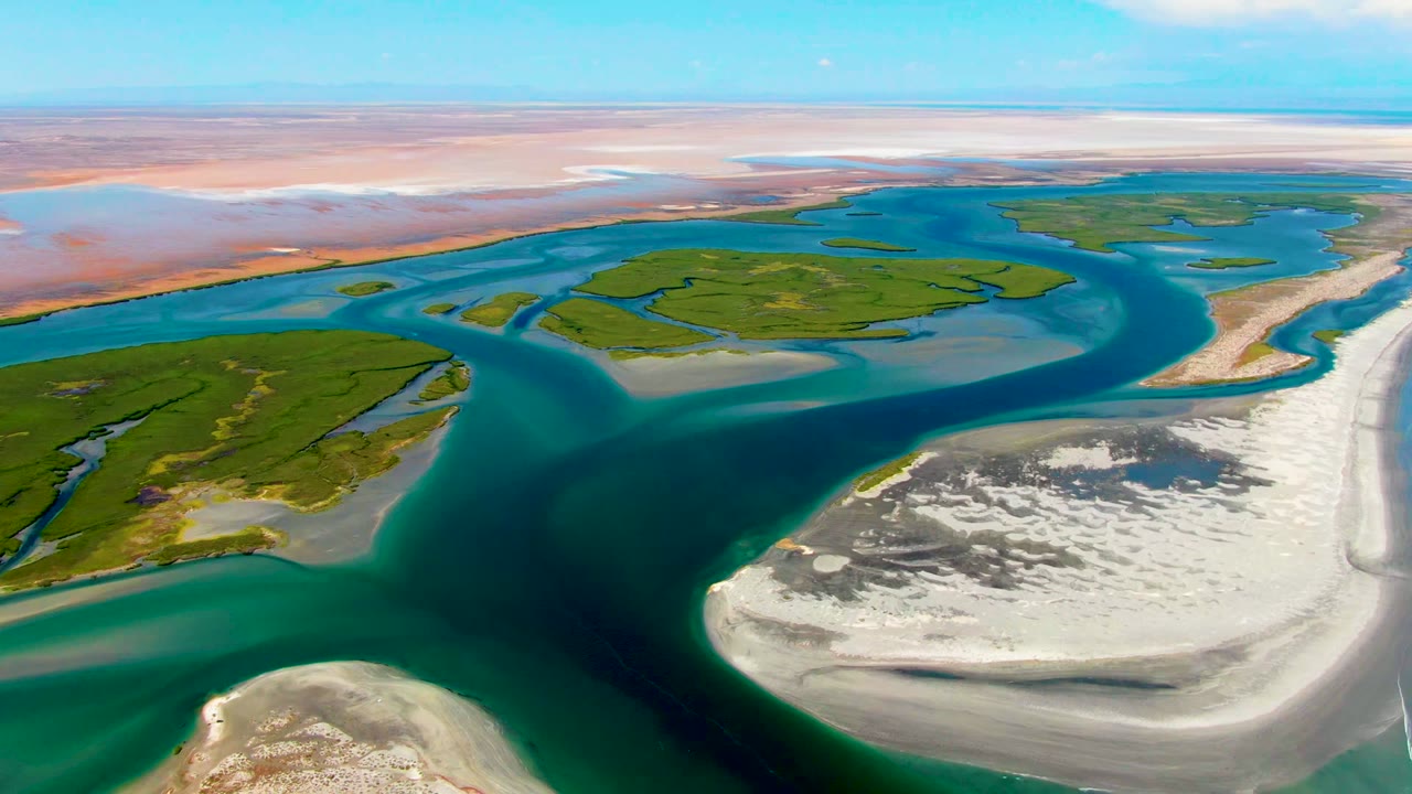 Wide drone view over La Bocana's delta pattern, with green island shapes bordered by blue water channels and bright sandy flats, and no visible people.