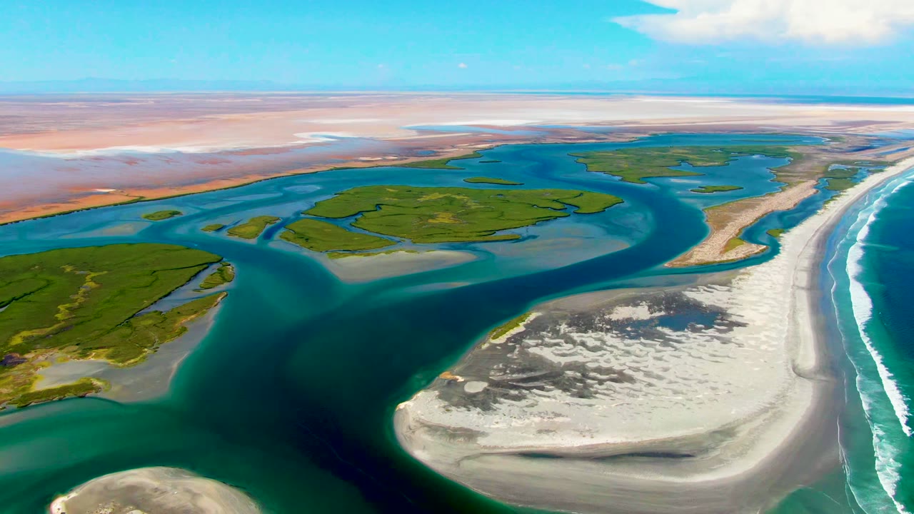 High aerial frame over La Bocana, Baja California Sur showing turquoise channels around white sandbars, green coastal land, surf on the right, pale flats on the horizon, and no visible people.